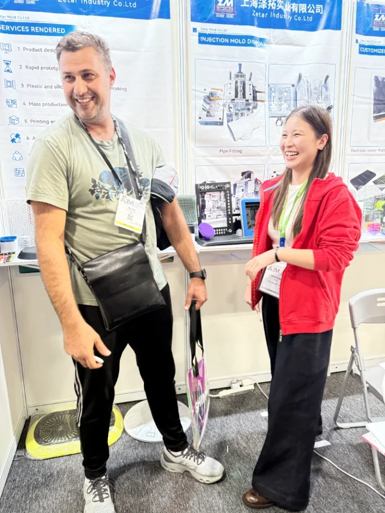 Two people talking at an industrial trade show booth with injection molding displays and product information in the background.