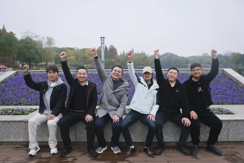 A group of seven people sit on a bench outdoors with raised fists, surrounded by purple flowers and trees.