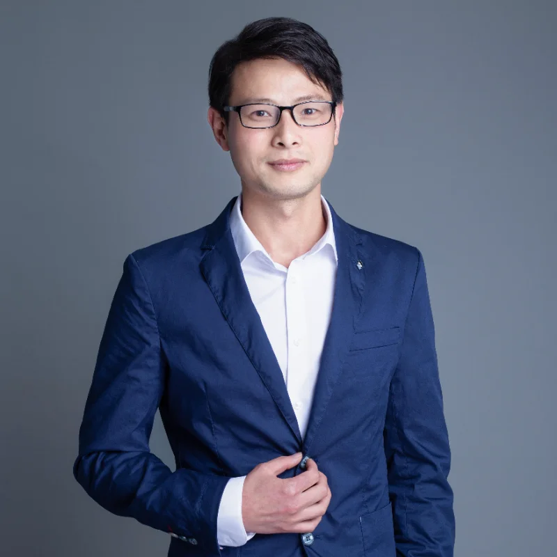 A person wearing a navy blue suit with a white shirt, standing against a gray background.