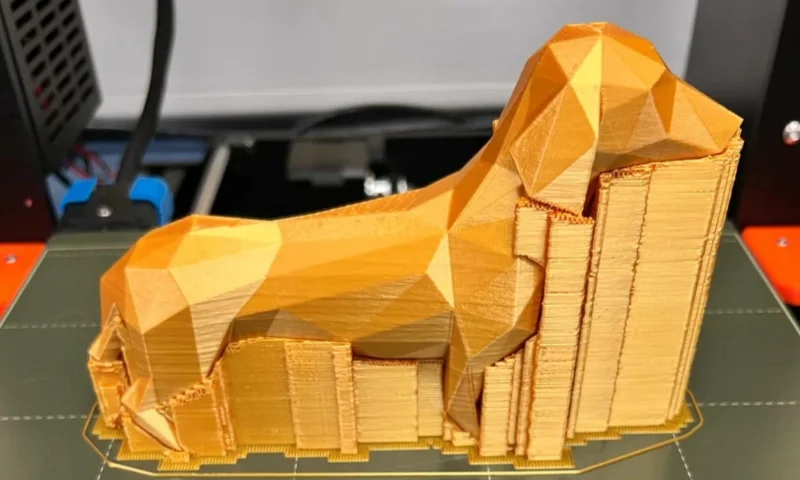 A gold-colored 3D printed dog model with geometric design and visible support structures on a printer bed.