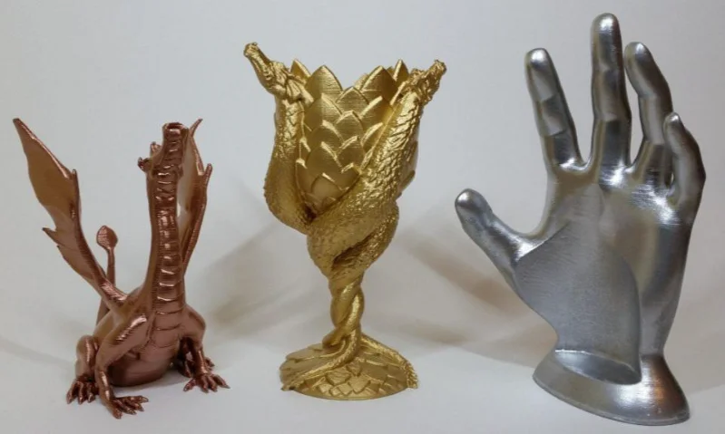 Three 3D printed objects: a bronze dragon, gold goblet, silver hand.