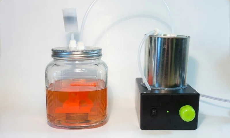 DIY anodizing setup with a glass container of orange liquid and connected metal device using tubes for the anodizing process.