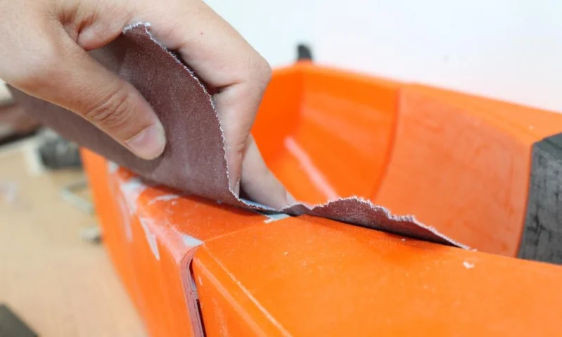 Hand holding sandpaper to smooth the edge of an orange plastic surface during finishing.